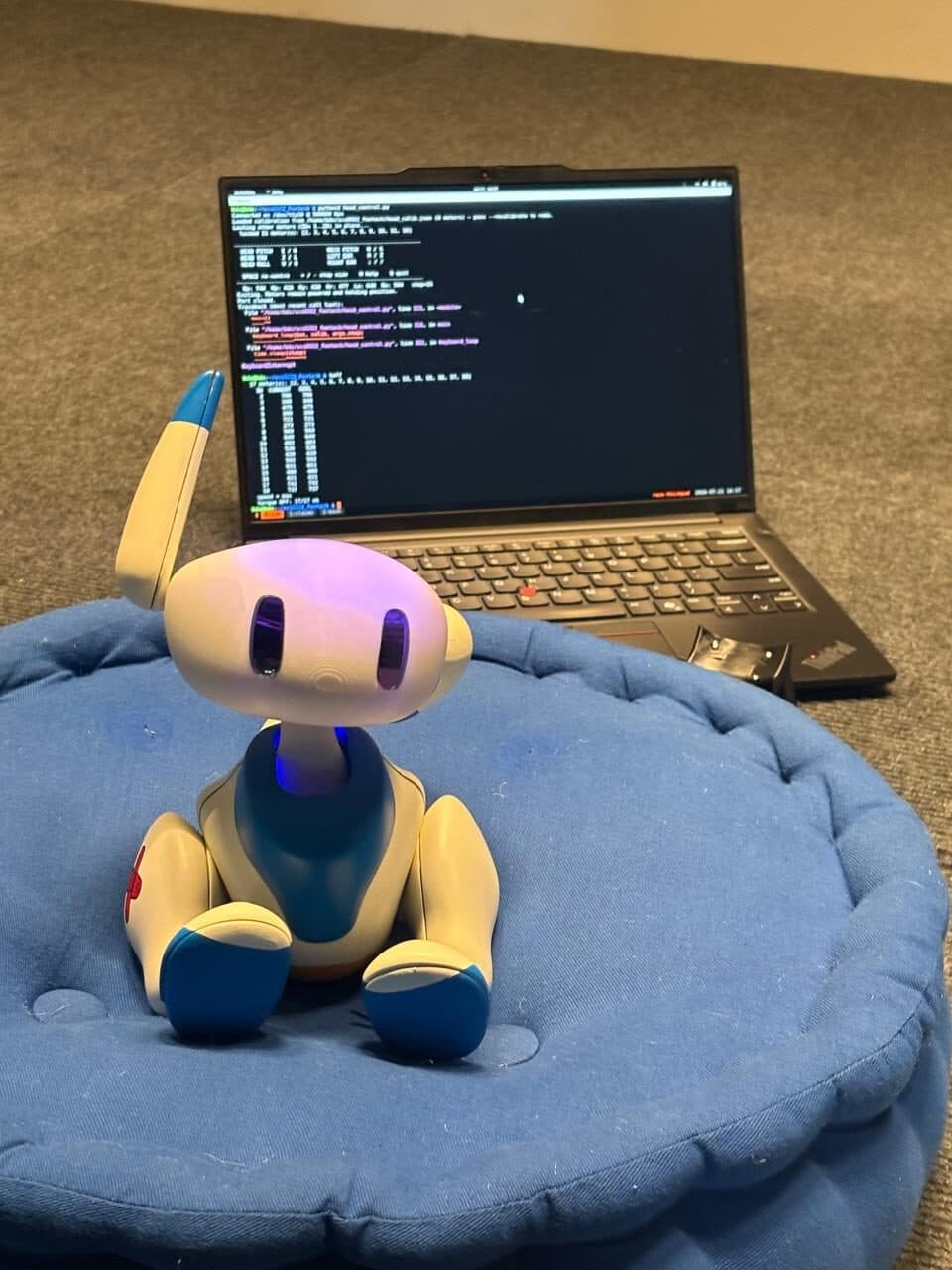 The same blue and white robot with one arm raised, sitting in front of a laptop showing a Python traceback in a terminal.