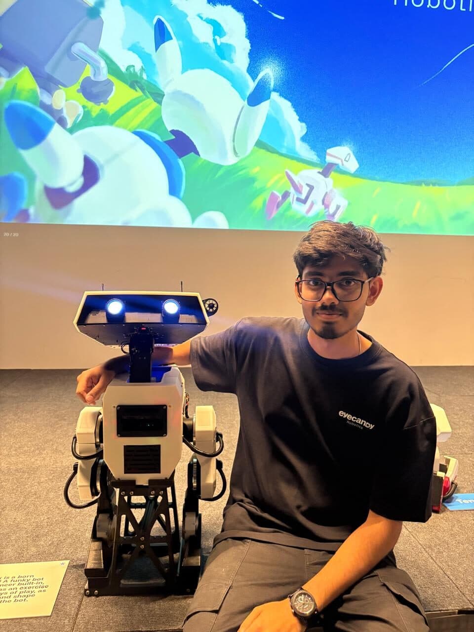 Kartikey Pathak kneeling beside a white humanoid robot at a public showcase, a projected slide behind him.